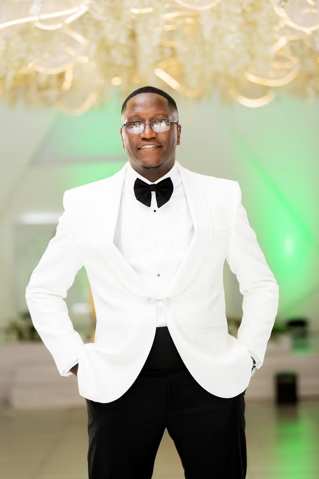 Portrait of Simba in a white dinner jacket beneath the lit floral chandelier