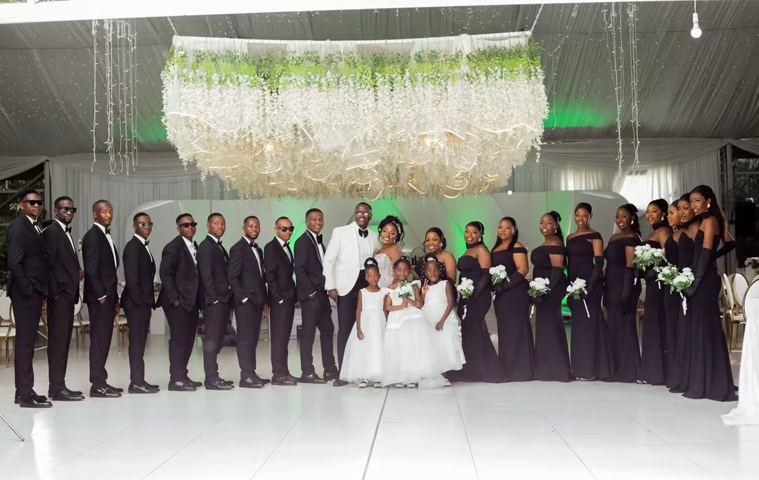 Full bridal party in black gowns and tuxedos under the floral chandelier